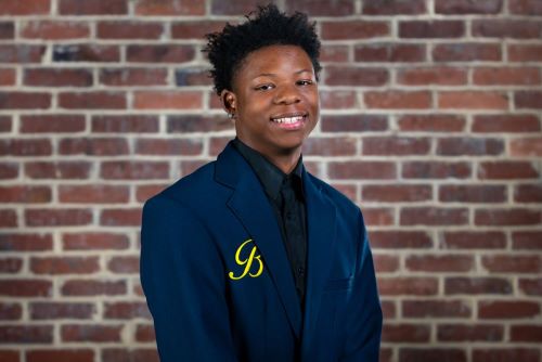 John "JT" Thompson, a young Black man, stands in front of brick background. He is wearing a navy blazer with the crest of Ballou's Academy of Hospitality and Tourism.