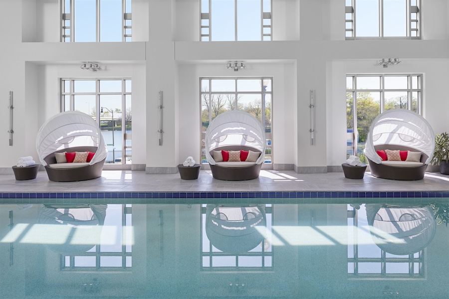 Indoor pool area with large windows, featuring three circular lounge chairs with canopies and red pillows, and small tables holding towels.