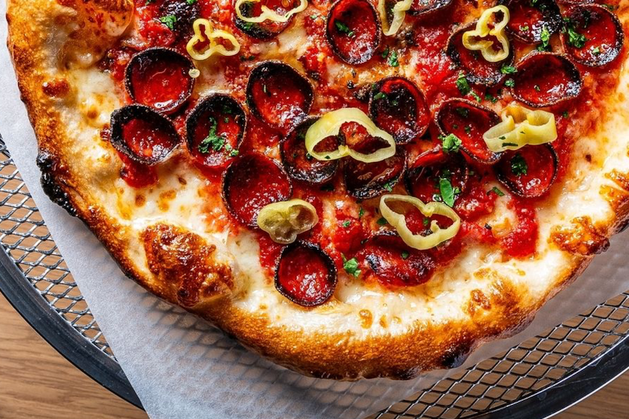 Close-up of a pepperoni pizza topped with melted cheese, red sauce, and sliced yellow peppers, served on a mesh pizza tray.