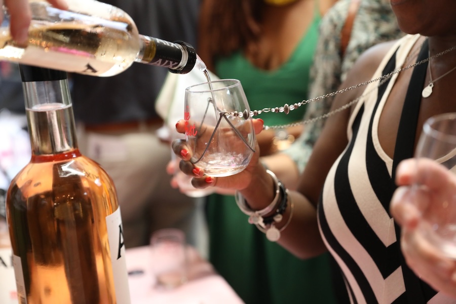 A close-up of a wine tasting event where a person is pouring wine from a bottle into a glass held by another person. The glass has a beaded necklace attached to it, and people are visible in the background.