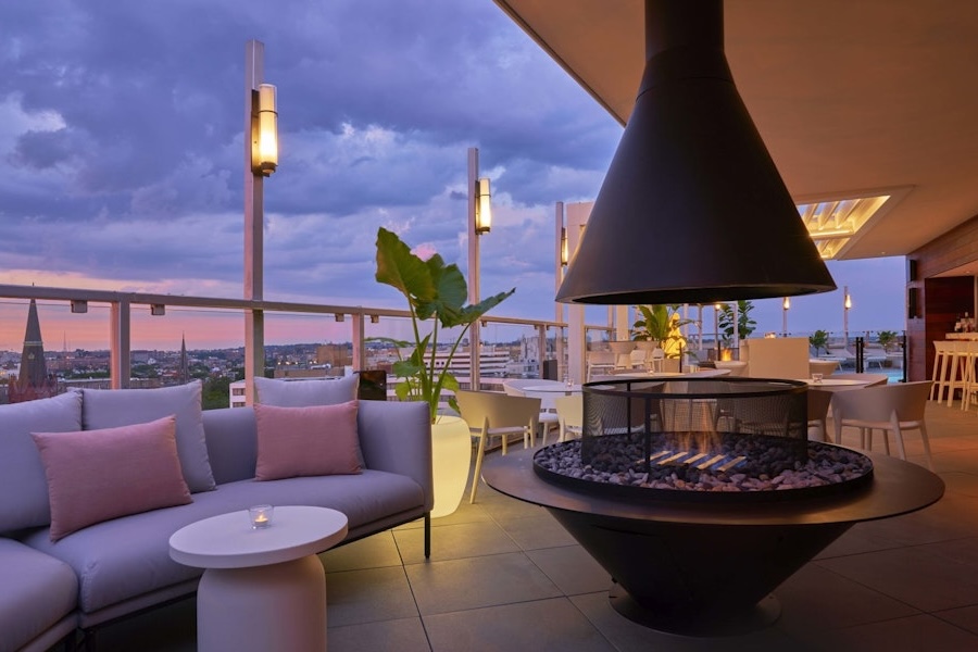 Rooftop lounge at sunset with modern furniture, including a sectional sofa with pink pillows, round tables, and a central fire pit. The area is illuminated with contemporary lighting and offers panoramic city views.