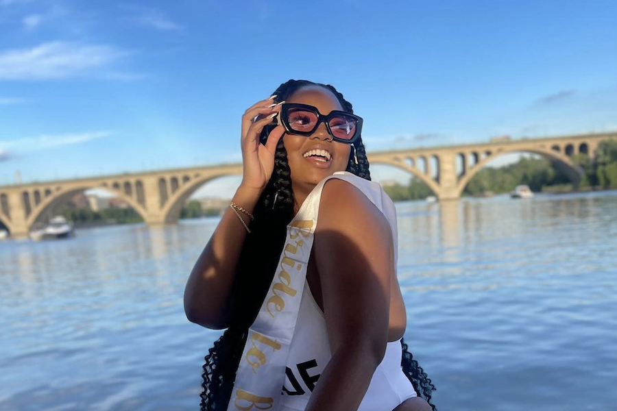 A smiling woman wearing large sunglasses and a 'Bride to Be' sash poses near a river with an arched bridge in the background.