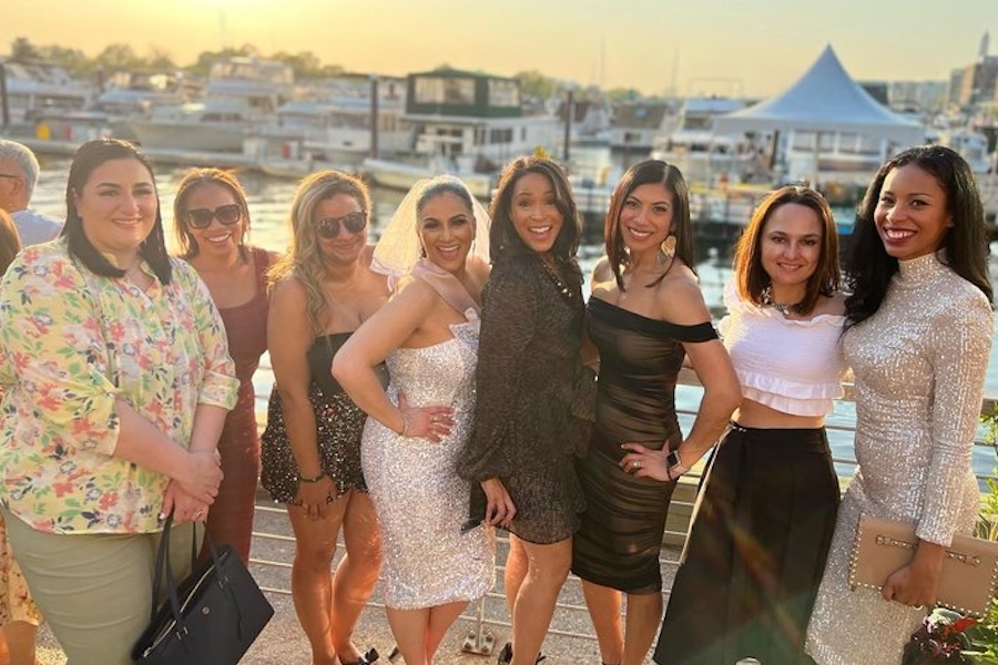 A group of women dressed in stylish outfits pose by the waterfront at sunset, with boats and a pavilion in the background.