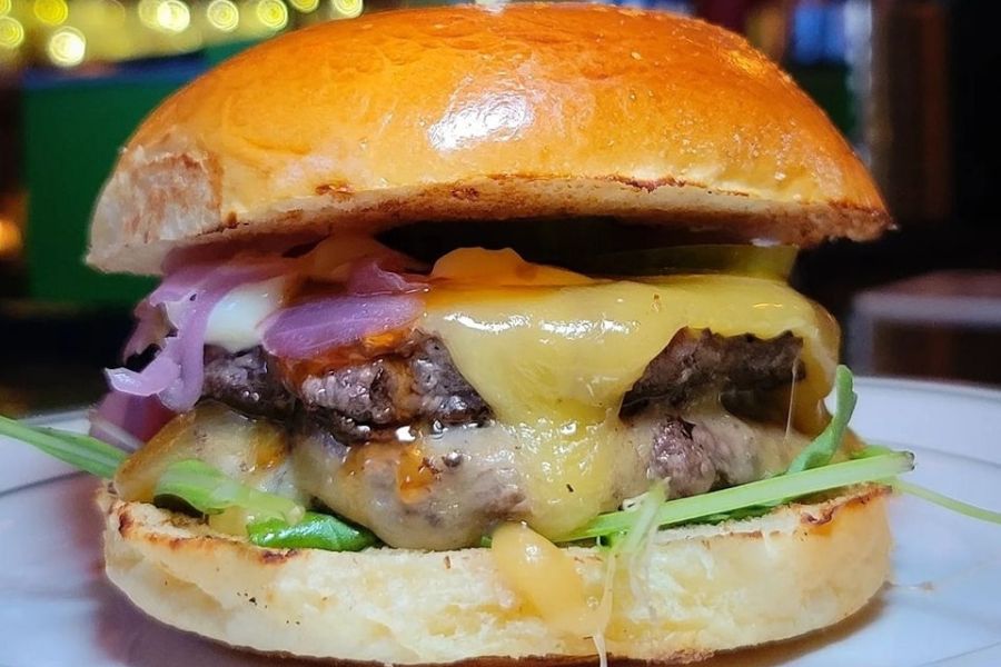 A close-up of the Proper Burger™ at Duke’s Grocery, featuring a juicy beef patty topped with melted cheese, onions and greens, served on a toasted bun.