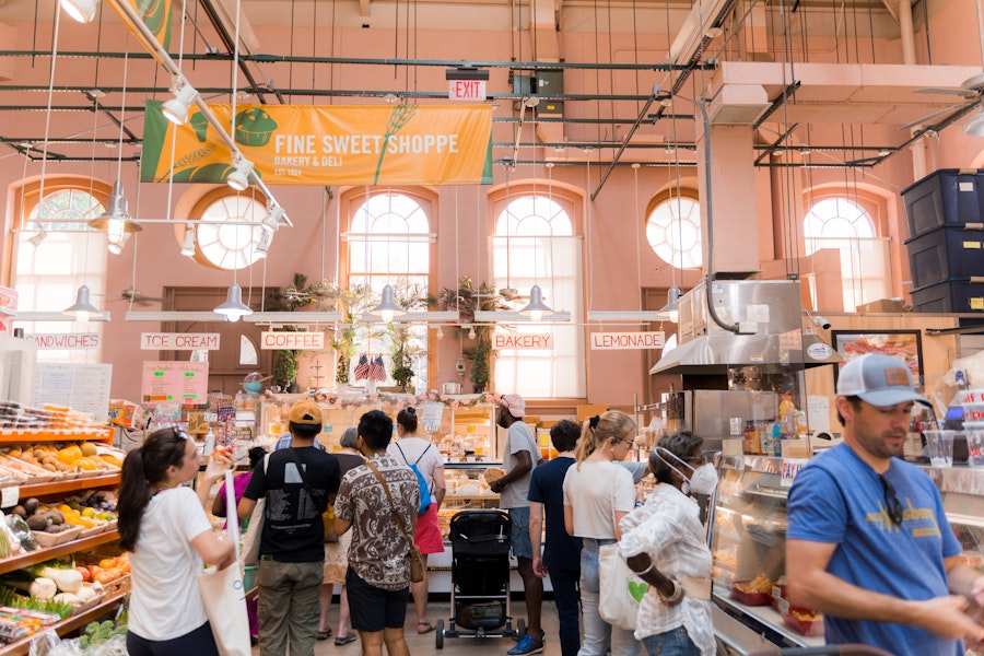 Shoppers line up at a bakery stall inside Eastern Market. The bright and airy space is filled with natural light, with signs for coffee, ice cream, and bakery items hanging above.