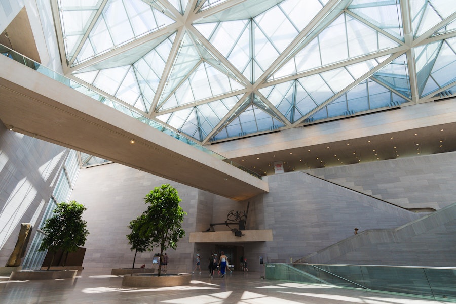 Interior view of a modern museum atrium with a glass ceiling, large open spaces, and minimalist architectural design, featuring a few visitors exploring the area.