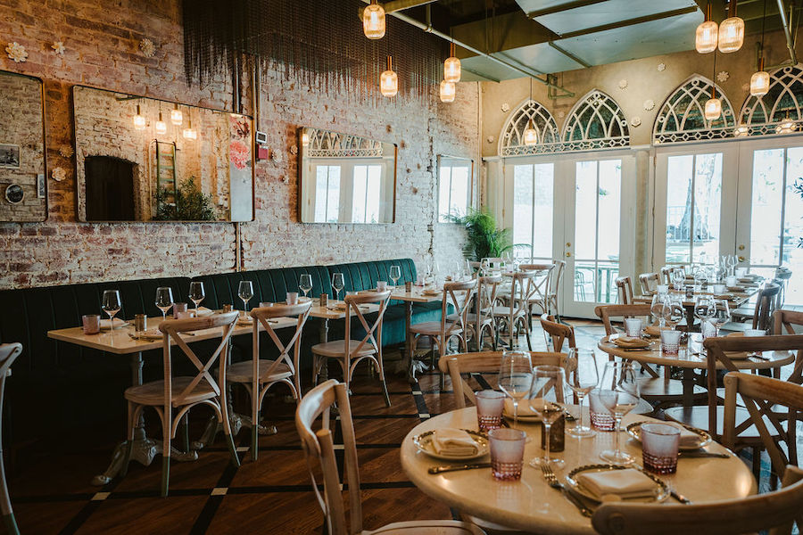 An elegant restaurant interior featuring exposed brick walls, arched windows, hanging lights, and neatly set tables with glassware and napkins.