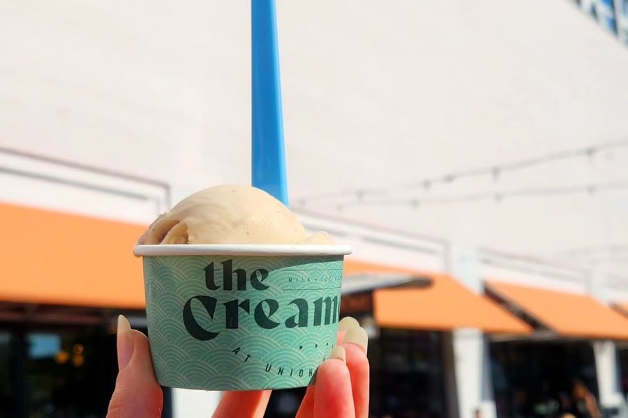 A hand holding a small cup of ice cream from The Creamery at Union Market with a bright blue spoon, against the backdrop of a building with orange awnings.