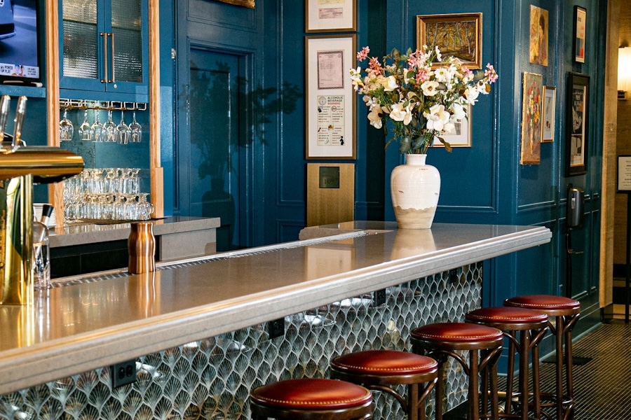 A stylish bar with rich blue walls, adorned with framed art, features a sleek counter lined with red leather stools and a vase of fresh flowers as a centerpiece.