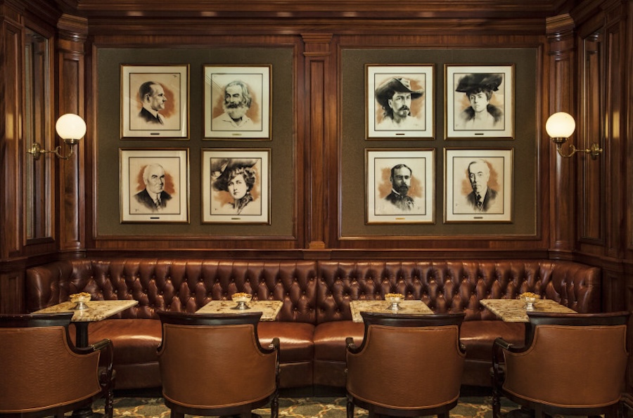 A cozy seating area featuring tufted leather banquettes, small marble tables, and portraits of notable historical figures adorning the walls in a warm wooden setting.