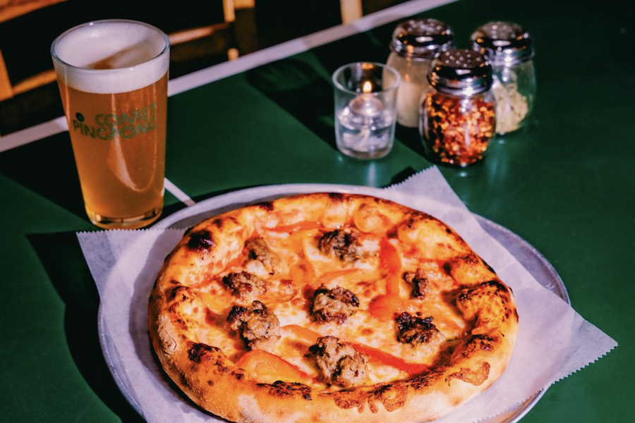 A delicious pizza topped with sausage sits on a round metal tray, accompanied by a tall glass of light beer. The table is green, and nearby condiments, including jars of crushed red pepper and a small candle, add to the inviting atmosphere.