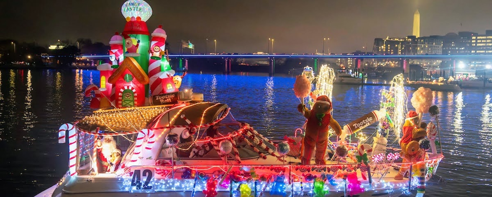 A festive boat decorated with candy cane and holiday lights glides along the water with the Washington Monument in the background.