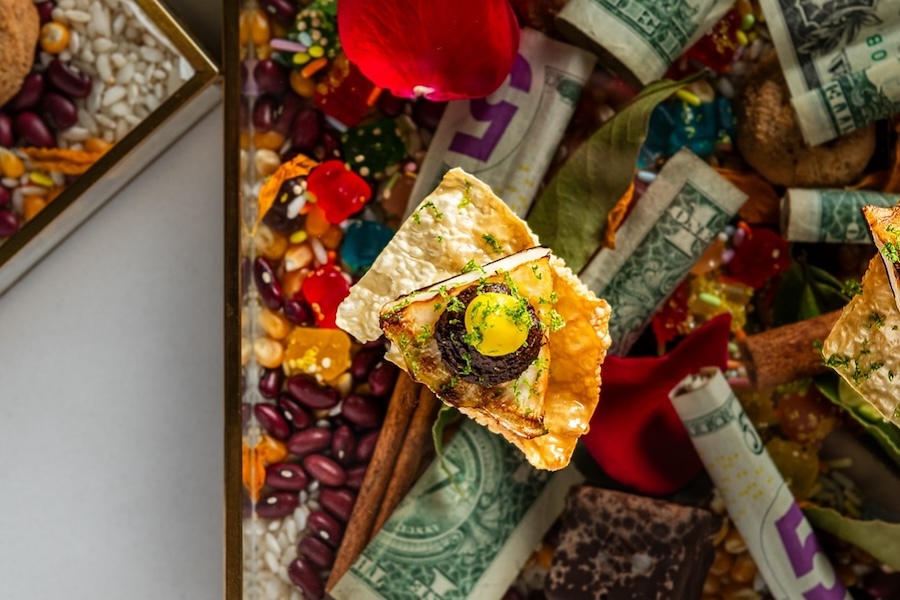 A close-up of an artistic dish surrounded by colorful ingredients, currency notes and decorative elements.