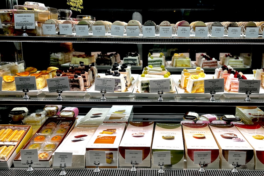 A glass bakery display case filled with colorful pastries, cakes, and macarons, each labeled with descriptions and prices.