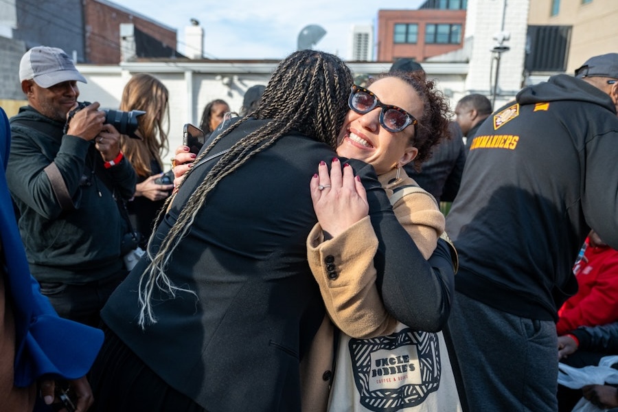 Two women embrace and smile during an outdoor go-go community event, surrounded by other attendees and photographers.