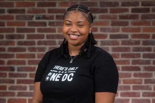 A smiling young Black woman stands against a brick wall with a There's Only 1 DC shirt