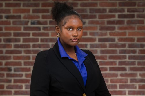 The professional headshot of Shamiyah, a young Black woman. She is wearing a black suit with a blue shirt and stands in front of a brick wall. 