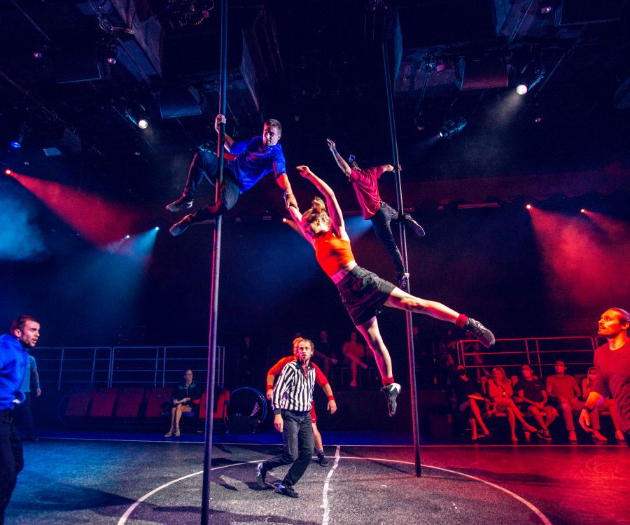 Acrobats perform daring moves on poles while a referee and audience watch during a high-energy scene from Duel Reality.