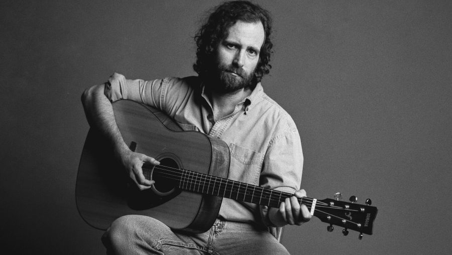 Comedian and musician Kyle Mooney poses with an acoustic guitar in a black and white promotional portrait.