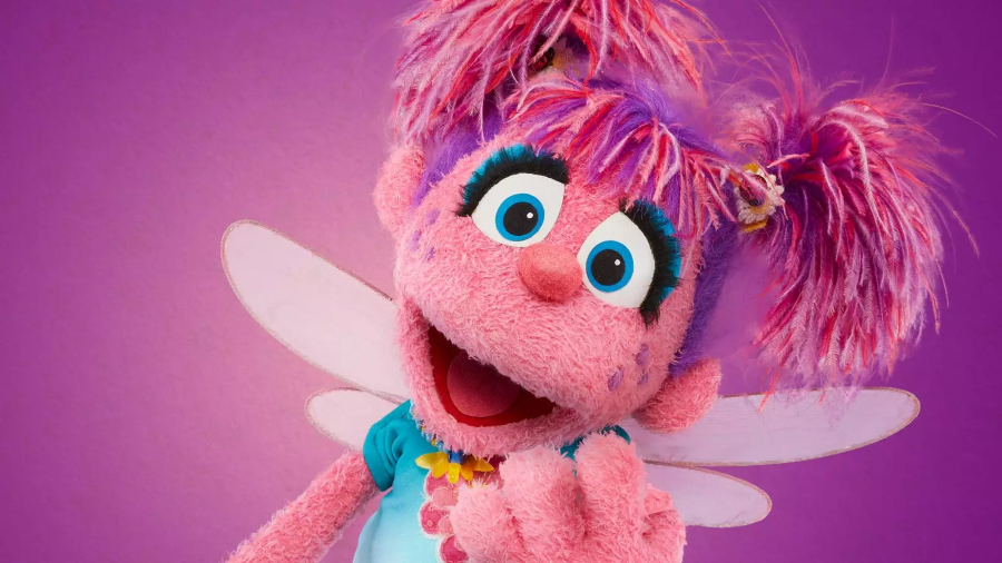 Abby Cadabby, a pink fairy Muppet from Sesame Street, poses joyfully against a vibrant purple background.