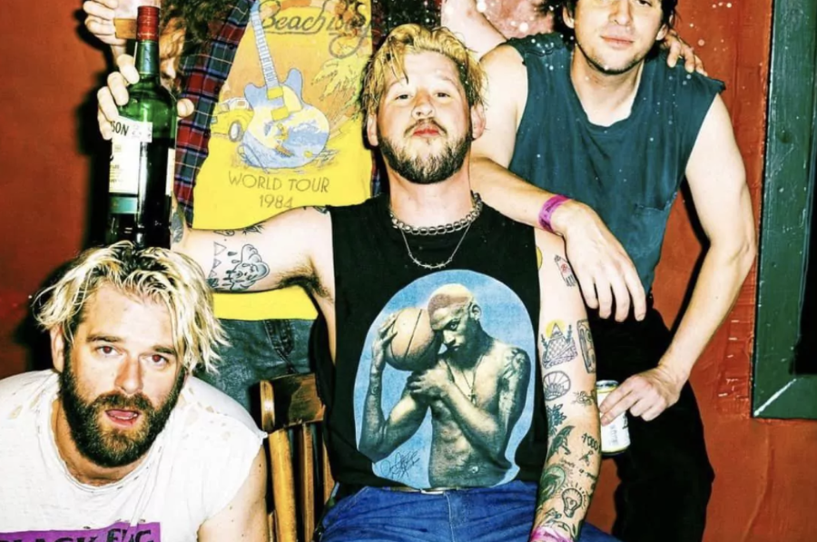 The band Wavves poses with a gritty, rebellious vibe. Members sport tattoos, drinks and rock-style attire in a candid indoor setting.