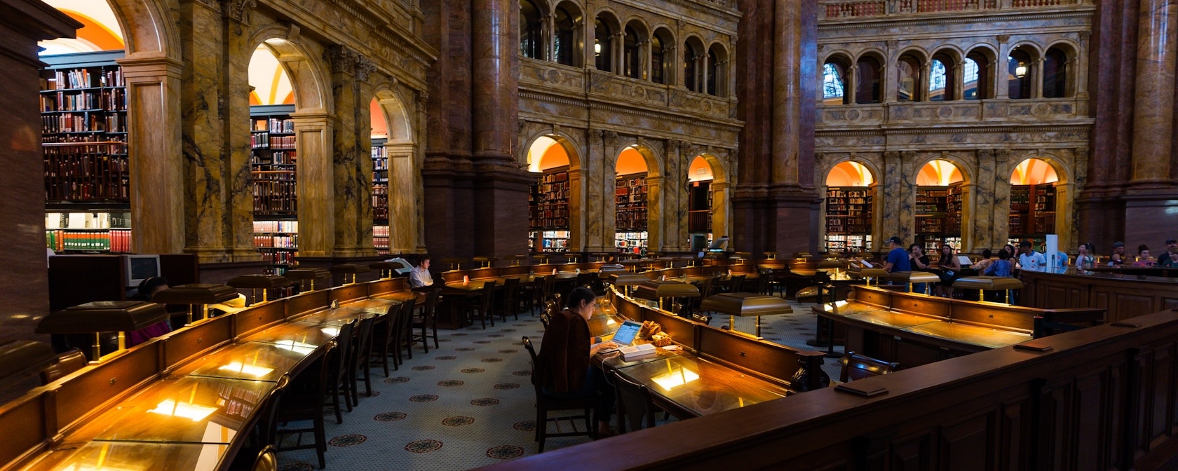 Visitors study and explore the grand marble reading room at the Library of Congress surrounded by arched alcoves and bookshelves.