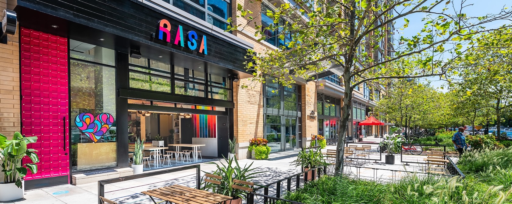 The exterior of Rasa restaurant in Mount Vernon Square features bright modern decor and a sunny outdoor patio with seating and greenery.