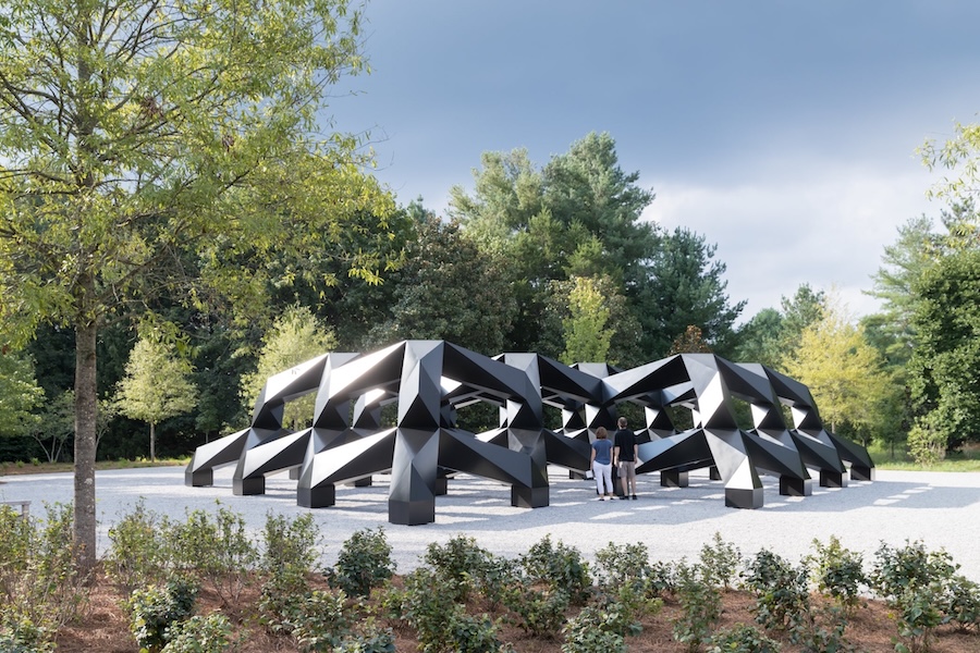 Tony Smith’s large black geometric sculpture “Smug” stands among trees and gravel at Glenstone Museum.