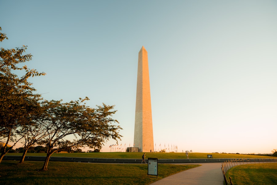 The Washington Monument rises above the National Mall as the sun sets and visitors walk along nearby paths.