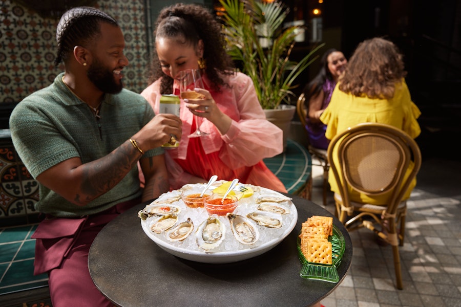 A couple clinks drinks at an outdoor restaurant table topped with oysters on ice and sauces.