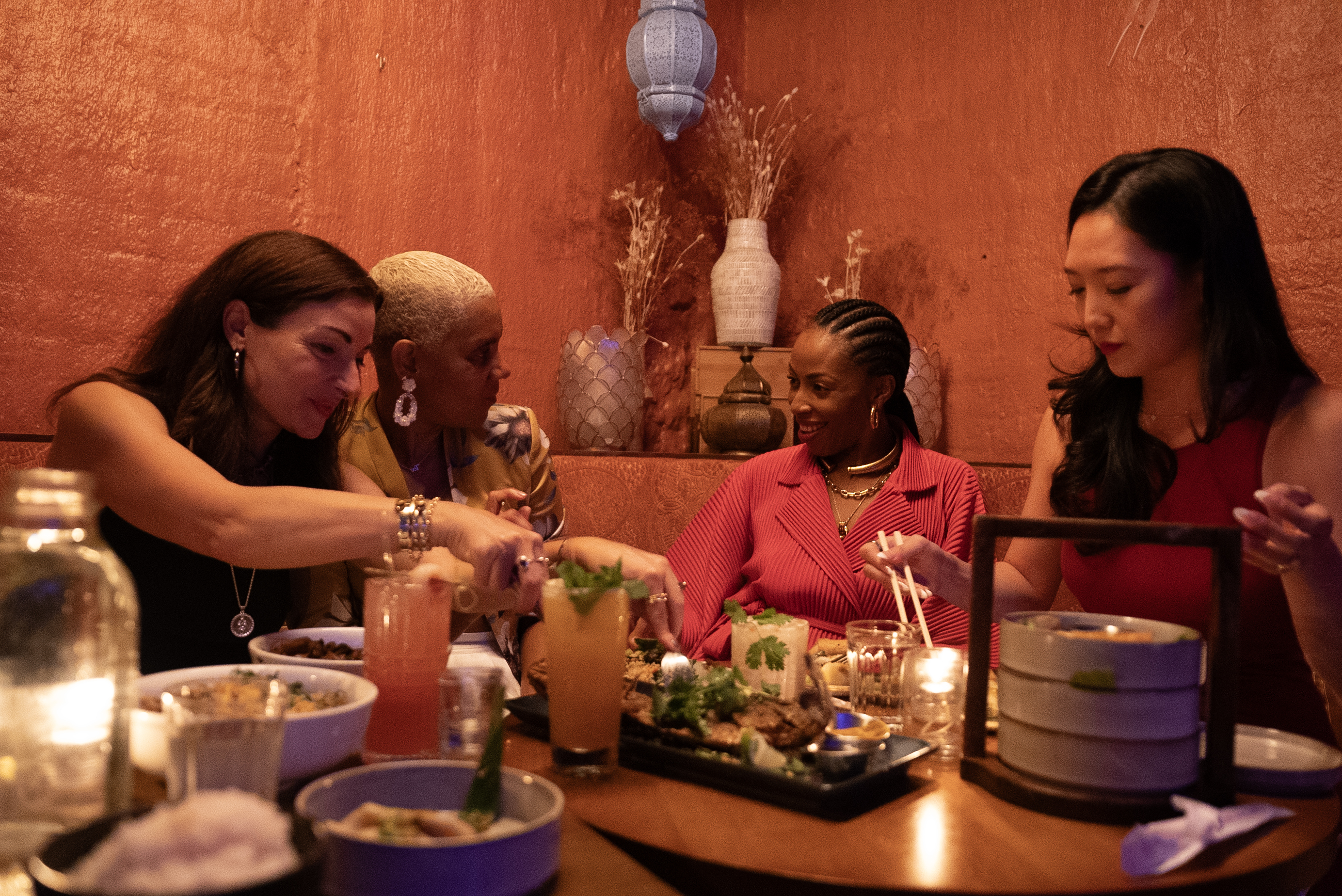 A group of friends share small plates and colorful cocktails at Doi Moi in Washington, DC, seated around a candlelit table with warm, textured walls in the background.