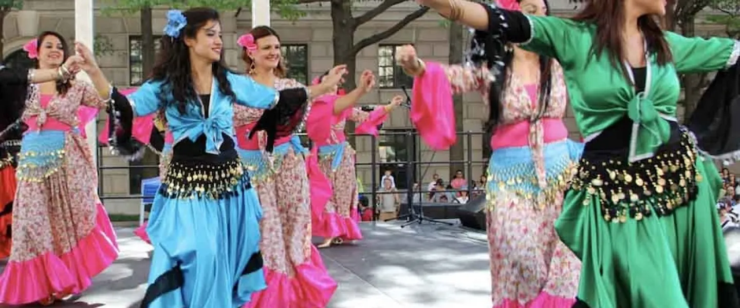 A group of women performing a Turkish dance on stage, wearing colorful traditional dresses with bright blue, green, and pink accents.