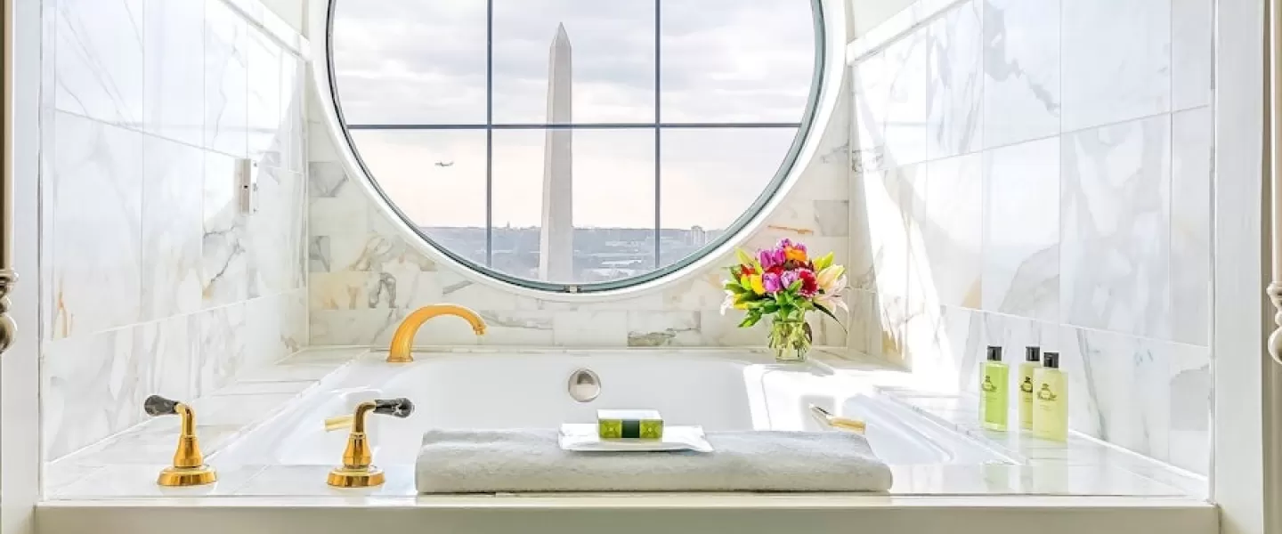A luxurious bathroom with a marble bathtub and golden fixtures, featuring a round window with a view of the Washington Monument. The bathtub has towels, flowers, and toiletries arranged neatly.