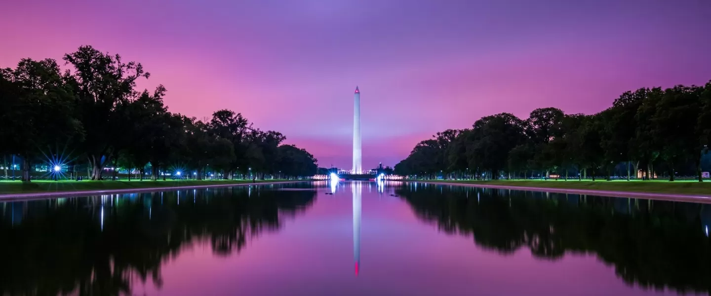 The Washington Monument reflects in the calm waters of the Lincoln Memorial Reflecting Pool at dusk, under a vibrant purple and pink sky.