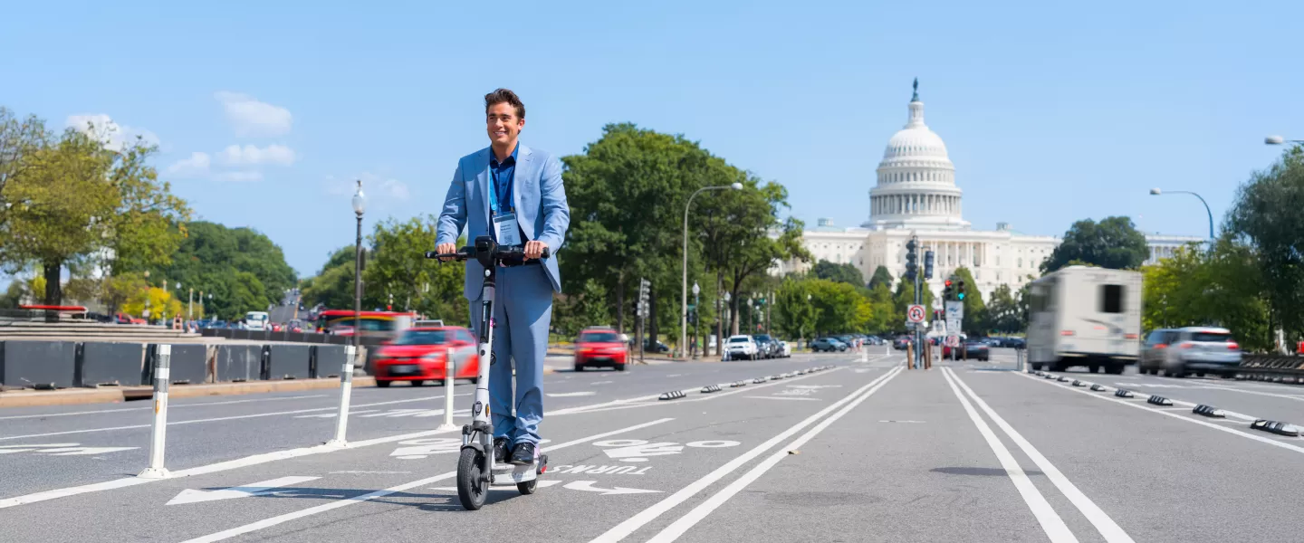 A man in a blue suit rides an electric scooter along a bike lane with the U.S. Capitol building in the background, enjoying a sunny day in Washington, DC.