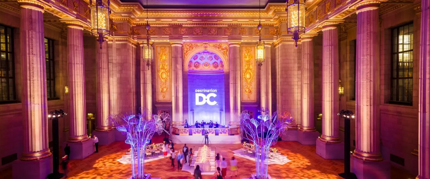 A festive crowd gathers in an ornate hall with tall columns, colorful lighting, and a large “DC” projection illuminating the space during a special event.