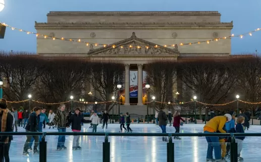 People ice skating at the National Gallery of Art Sculpture Garden rink during the evening, with string lights overhead and the museum building in the background.