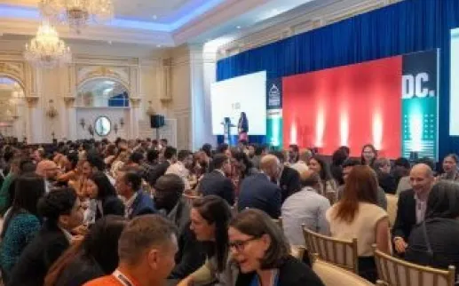 A lively networking session in a grand ballroom with chandeliers, where rows of attendees sit face-to-face engaging in timed conversations while a speaker stands on stage in front of a large red and blue backdrop.
