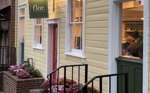 The exterior of Flor Coffee + Books, a quaint home painted yellow with inviting flowers and a red door. 

