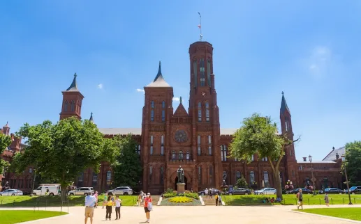 Visitors walk across the National Mall toward the red sandstone Smithsonian Castle on a sunny day.
