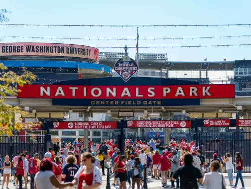 Crowds of fans dressed in red gather at the Center Field Gate of Nationals Park in Washington, DC, with a large "2019 World Champions" sign above the entrance.