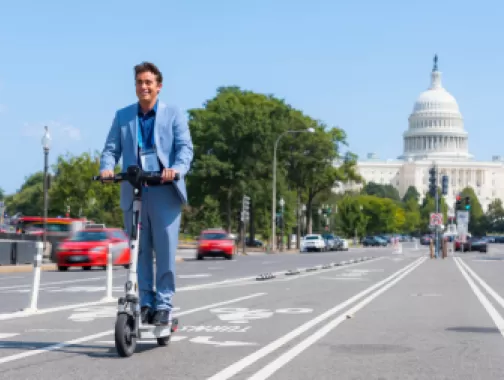 A man in a blue suit rides an electric scooter along a bike lane with the U.S. Capitol building in the background, enjoying a sunny day in Washington, DC.