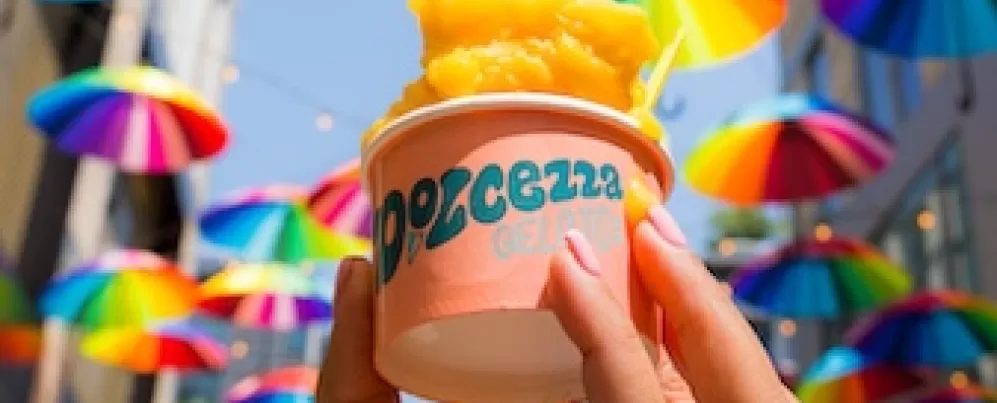 A hand holding a cup of orange gelato with vibrant, rainbow-colored umbrellas hanging overhead in the background.