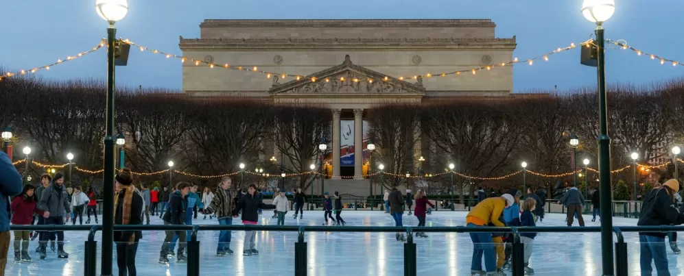 People ice skating at the National Gallery of Art Sculpture Garden rink during the evening, with string lights overhead and the museum building in the background.