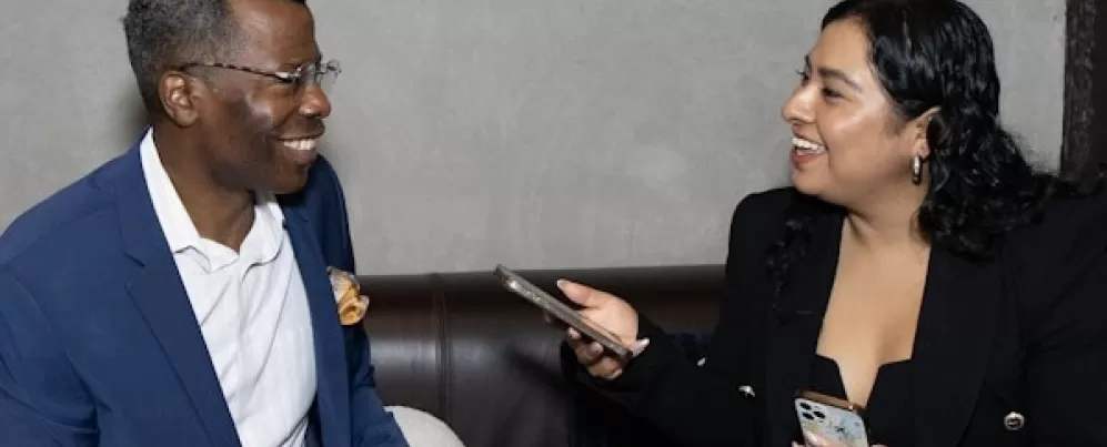 This image shows two people sitting on a couch, engaged in a friendly and professional conversation. The woman on the right is holding a phone, possibly conducting an interview. Both are smiling, suggesting a positive and engaging discussion. The setting appears casual yet professional, possibly a media or networking event.