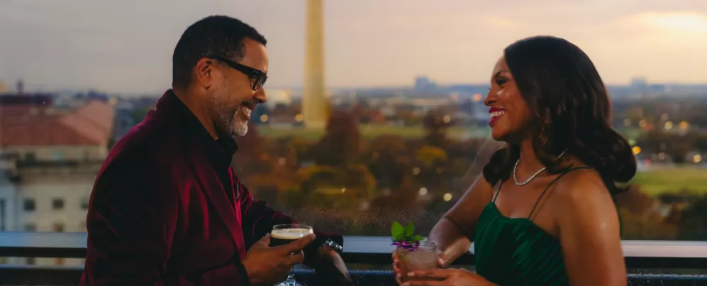A well-dressed couple enjoys cocktails at The Vue rooftop restaurant with the Washington Monument in the background at sunset in Washington, DC.