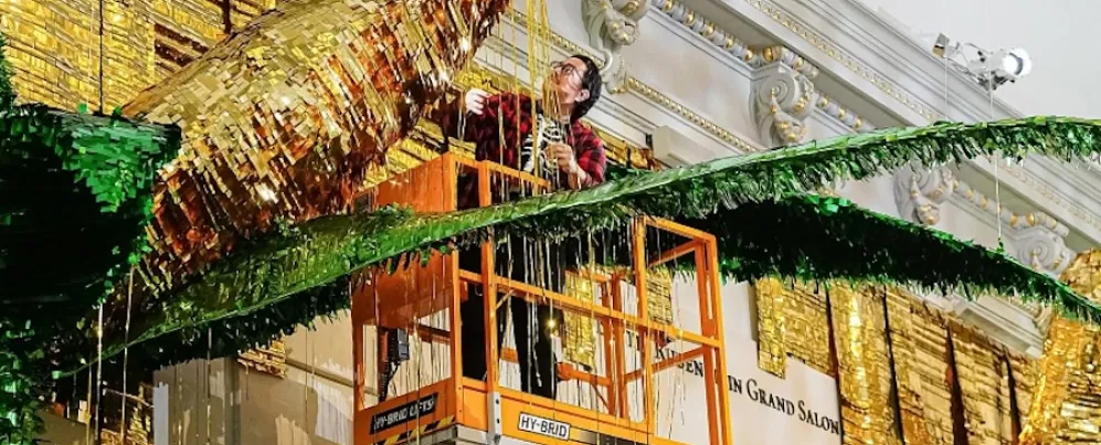 Artist installs a large gold and green piñata-style artwork from a lift inside a grand gallery.