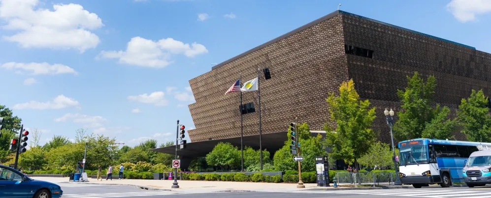 A view of the National Museum of African American History and Culture on a sunny day as cars pass by. . 