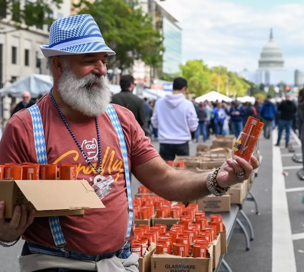 A man with a white beard and a blue checkered hat and suspenders, handing out orange beer cans at an Oktoberfest event in Washington D.C., with the U.S. Capitol building visible in the background.